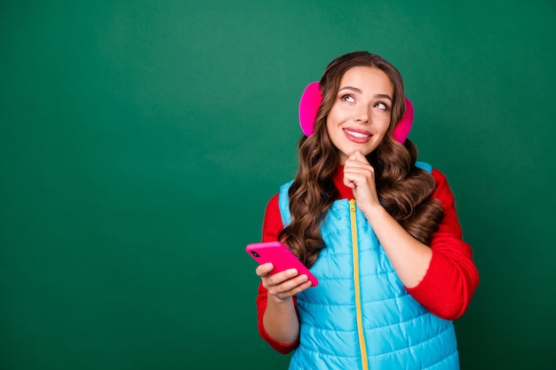 Photo of charming cute young lady look empty space finger chin think news choosing best shopping option wear pink ear warmers blue zipped vest red pullover isolated green color background