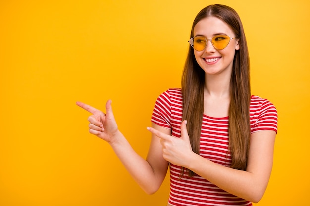 Photo of charming lovely young girl smiling pointing fingers empty space suggestion recommendation best variant wear sun specs striped white red shirt vibrant yellow color background