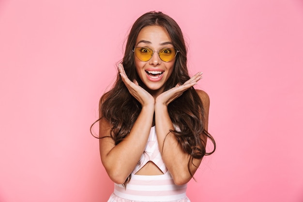 Photo of charming woman 20s wearing sunglasses smiling and rejoicing, isolated over pink wall