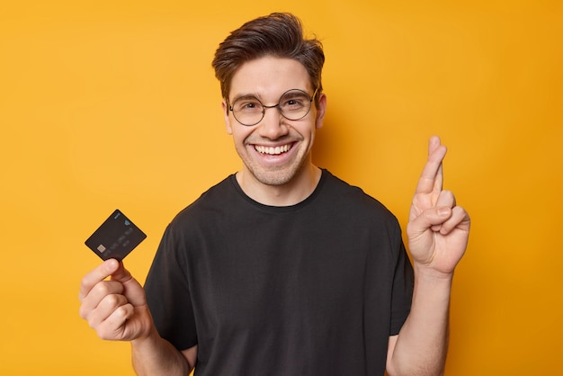 Photo of cheerful adult man keeps finger crossed believes in good luck holds credit card makes wish to recieve lump sum of money on his bank account wears round spectacles casual black t shirt