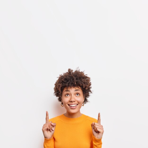 Photo of cheerful African American female points fingers up at top promo text suggests to check out awesome offer demonstrates copy space banner against white wall wears casual jumper.