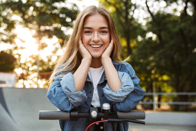 Photo of a cheerful beautiful happy young teenage girl outside in nature green park walking with scooter.