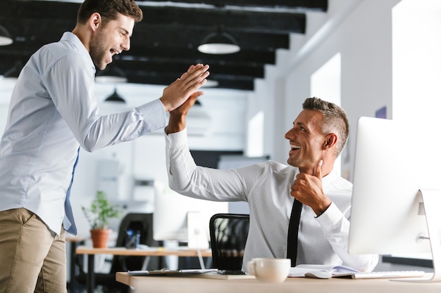 Photo photo of cheerful businessmen 30s in formal clothes giving high five together in office, during successful deal