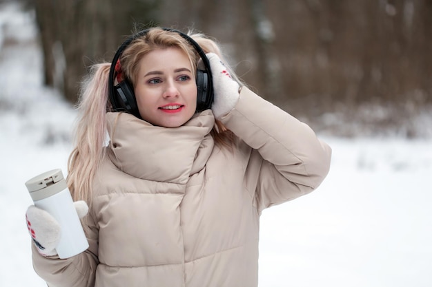 Foto di una giovane ragazza sorridente allegra e felice con le cuffie e una tazza termica con tè caldo in inverno