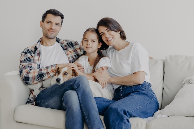 Photo of cheerful husband and wife spend free time with their\
lovely daughter and favourite domestic animal sit all together at\
sofa have good mood enjoy togetherness show love to each other