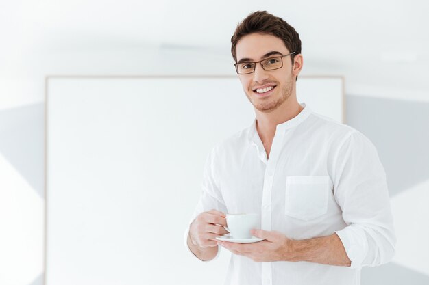 Photo of cheerful man wearing eyeglasses and dressed in white shirt holding cup of coffee near big board. Look at camera.