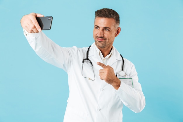 Фотография жизнерадостного врача в белом халате и стетоскопа, делающего селфи на мобильном телефоне, стоящего изолированно над синей стеной