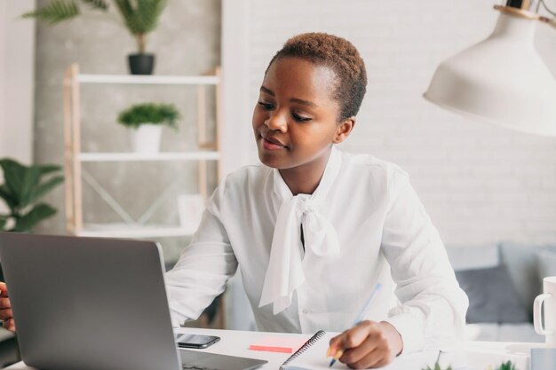 Photo of cheerful mixedrace woman in white shirt working with laptop while sitting at table in office Teamwork business meeting Freelancers working Student learning online