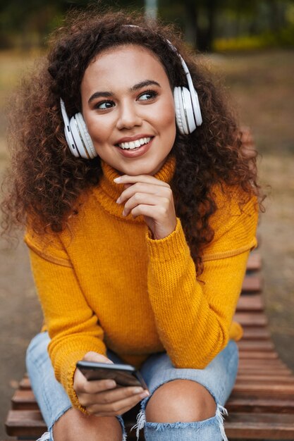 Photo of a cheerful optimistic beautiful young curly woman sit on bench in park outdoors listening music with headphones using mobile phone.