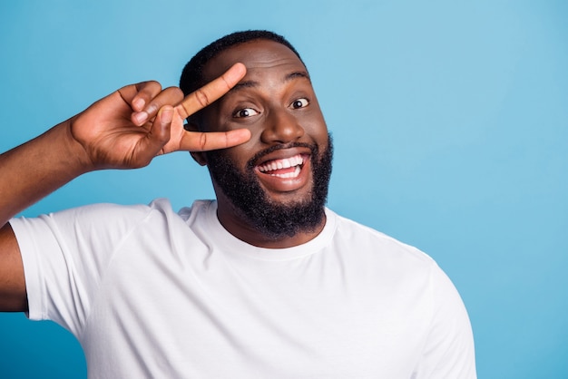 Photo of cheerful positive guy shiny smile show v-sign cover eye on blue background