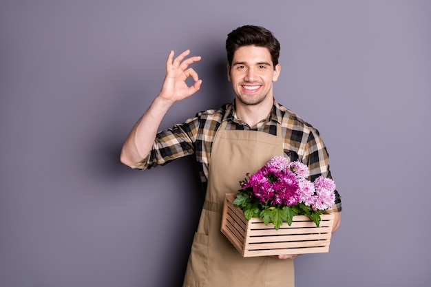 Foto di allegro positivo toothy raggiante uomo sorridente a trentadue denti mostrando segno ok che tiene la scatola di fiori con la mano isolata di colore grigio parete