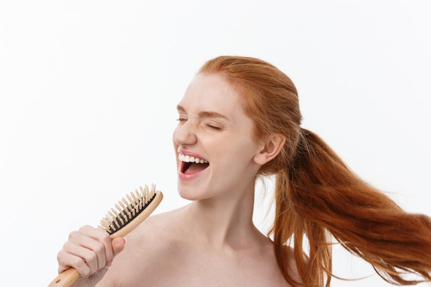 Photo of cheerful smiling freckled ginger young woman combs her long red hair, glad to prepare for date with boyfriend, isolated over white