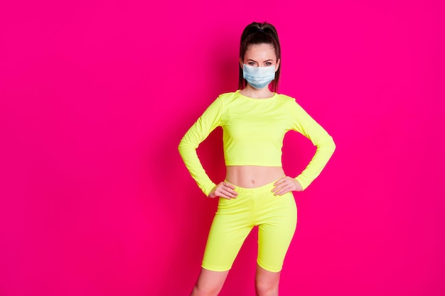 Photo of cheerful sweet young woman dressed sportwear mask hands arms waist isolated pink color background