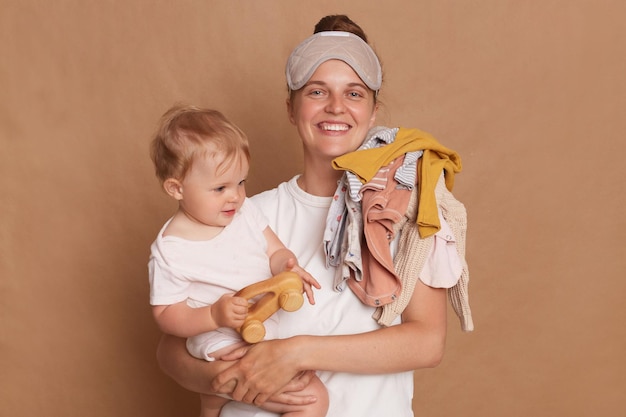 Photo of cheerful woman wearing white T shirt and blindfold holding her baby daughter in hands isolated over brown background expressing happiness enjoying maternity leave