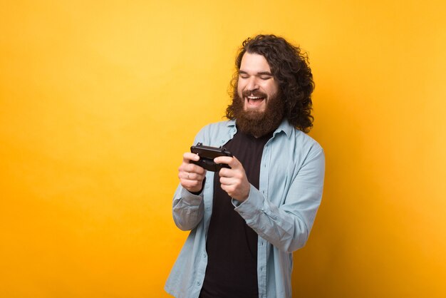 黄色の背景の上のスマートフォンでゲームをしている陽気な若いひげを生やした男の写真