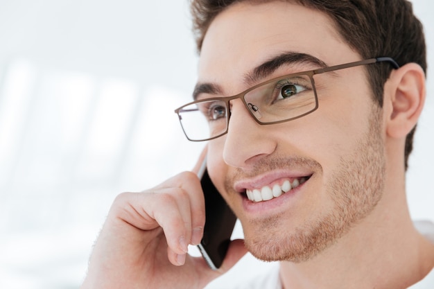 Photo of cheerful young man dressed in white shirt standing near big white window while talking by phone. Look aside.