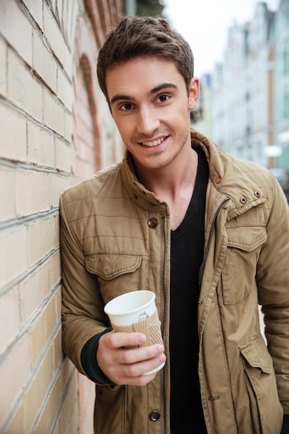 Photo of cheerful young man walking on the street and looking at camera while holding cup of coffee.