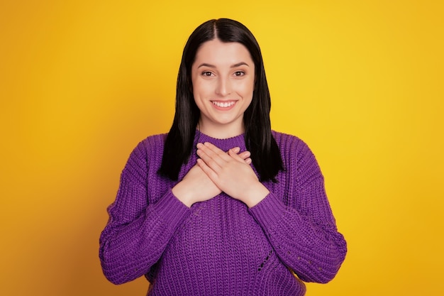 Photo of cheerful young woman keeps hands on chest demonstrates sign of gratitude and affectionisolated over yellow studio background