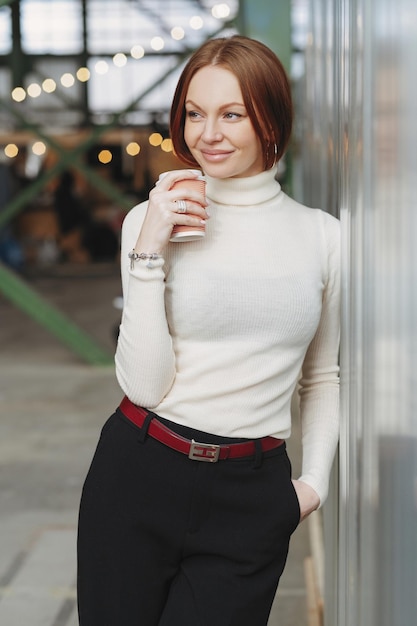 Photo of cheerful young woman wears elegant clothes dressed in white jumper and black trousers drinks disposable cup of coffee dreams about something pleasant keeps gaze aside Rest concept