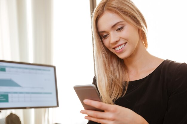 Photo of cheerful young woman worker sitting in office near computer while chatting by phone. Look at phone.