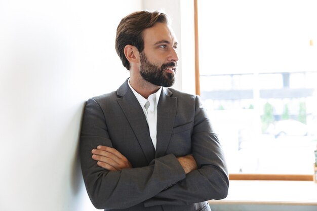 Foto foto di un allegro uomo d'affari barbuto positivo in posa in ufficio vicino alla finestra al chiuso con le braccia incrociate.