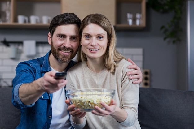 Фотография близкой пары мужчины и женщины вместе дома, весело и с удовольствием смотрящих телевизор и поедающих попкорн