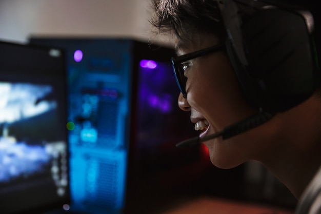 Photo closeup of asian pleased gamer boy playing video games online on computer in dark room, wearing headphones with microphone and using backlit colorful keyboard