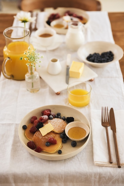 Primo piano fotografico di piatti e posate per la colazione. pancake con frutti di bosco, latte, succo d'arancia, caffè sul tavolo della cucina in appartamento al mattino