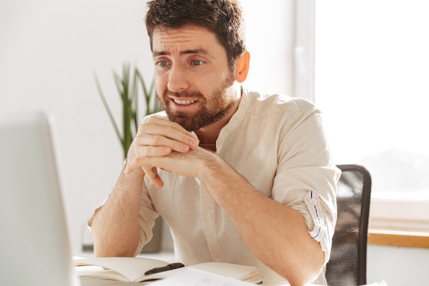 Photo closeup of european businessman 30s wearing white shirt using laptop, while sitting at table in modern office