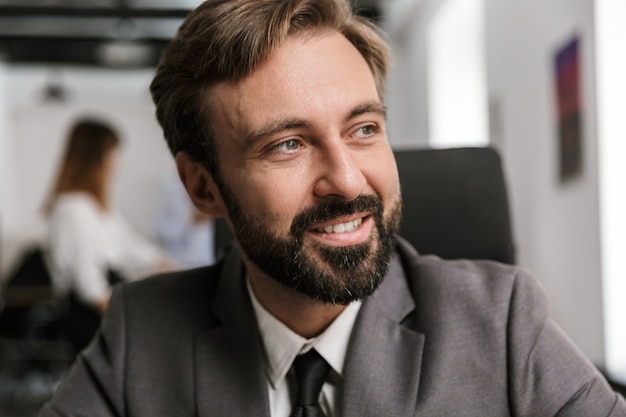 Крупным планом фото счастливого бизнесмена в официальном костюме, улыбающегося и смотрящего в сторону, сидя за столом в офисе открытой планировки
