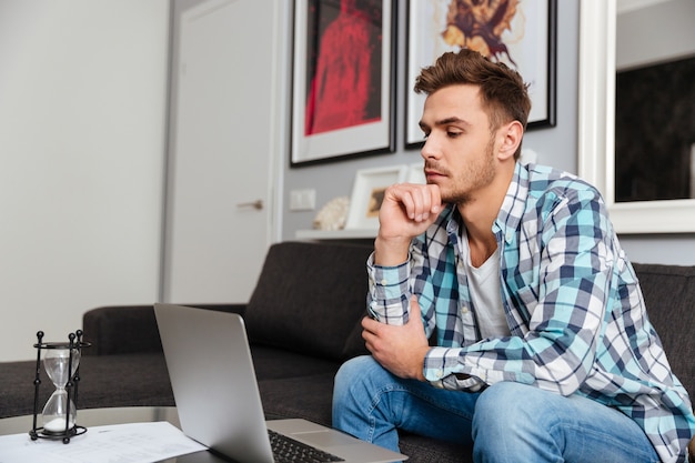 Photo of concentrated bristle man dressed in shirt in a cage print sitting on sofa in home and using laptop computer