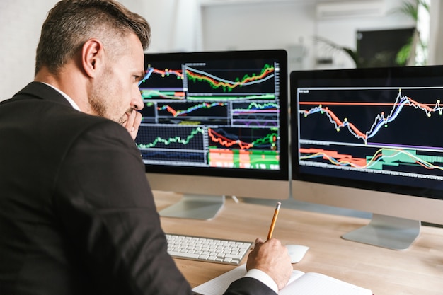 Photo of concentrated businessman 30s wearing suit sitting at table in office and working with graphics and charts on computer