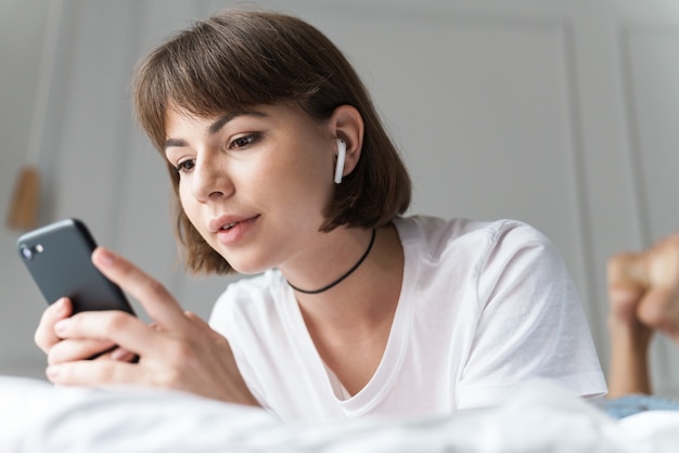 Photo of a concentrated young pretty woman indoors at home listening music with bluetooth earphones using mobile phone.