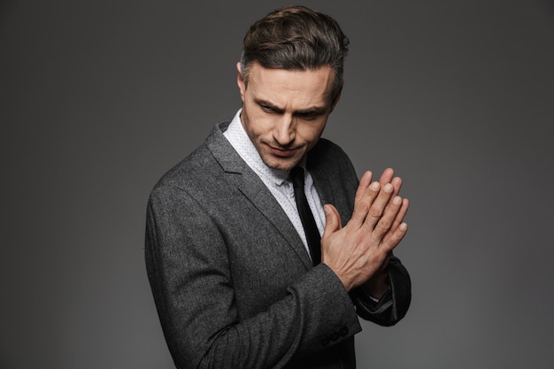 Photo of confident business man 30s wearing formal suit and black tie looking downward holding hands together, isolated over gray wall