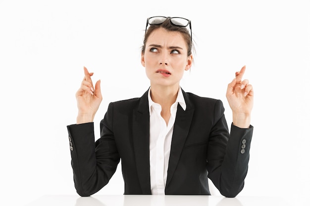 Photo of confident female worker businesswoman dressed in formal wear keeping fingers crossed while working and sitting at desk in office isolated over white wall