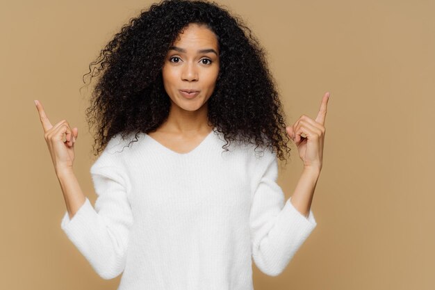 Photo of confident woman has bushy Afro hairstyle points both index fingers above dressed in white jumper stands against brown background advertises some item shows direction Come upstairs