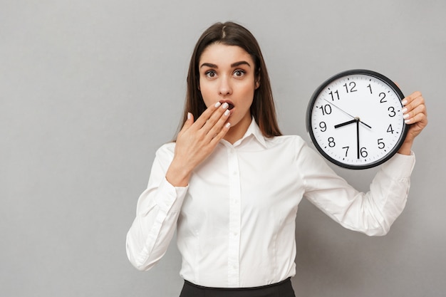 Photo of confused office woman in white shirt and black skirt holding big round clock and being concerned about time, isolated over gray wall