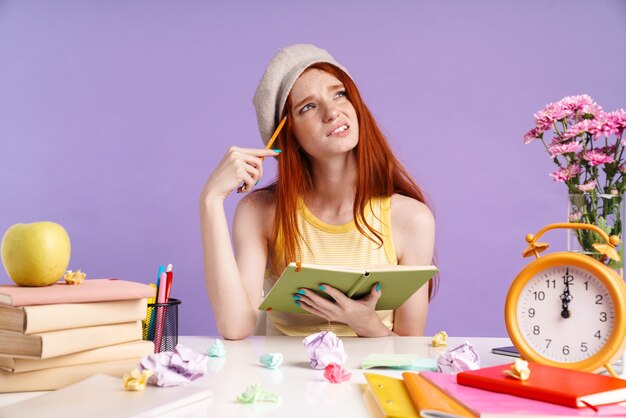 Photo photo of confused student girl writing in exercise book while doing homework isolated over purple wall