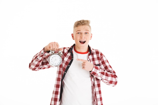 Photo of confused teen man  wearing plaid shirt holding alarm and pointing finger on clock face, isolated over white wall