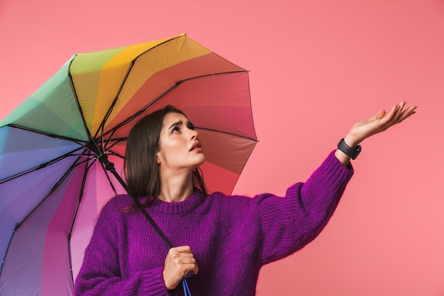 Photo of a confused young woman posing isolated over pink space holding rainbow umbrella.