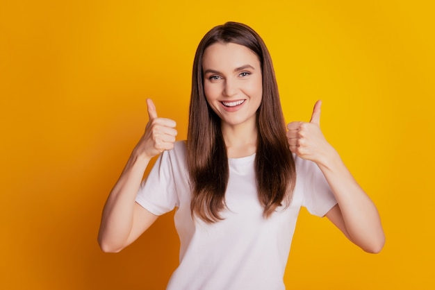 Photo of cool cheerful lady raise two thumbs up toothy smile wear white t-shirt posing on yellow background