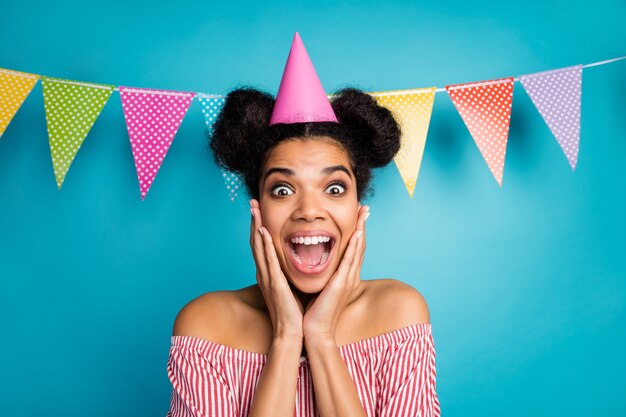 Photo of crazy dark skin lady arms cheekbones unexpected surprise birthday party wear cone cap red white striped shirt naked shoulders colorful dotted flags hang over blue color wall