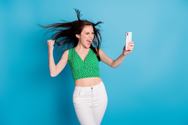 Фотография сумасшедшей восторженной девушки, пользующейся мобильным телефоном, празднует подписчики, цель, победа, поднять кулаки, кричать, да, носить белые брюки, брюки, майку, изолированную на синем цветном фоне