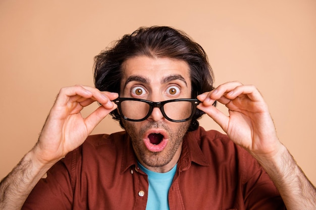 Photo of crazy surprised young man fingers hold glasses open mouth wear spectacles brown t-shirt isolated beige color background