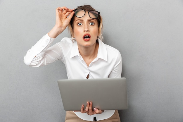 Photo of curious businesslike woman wearing glasses standing and holding laptop in the office, isolated 