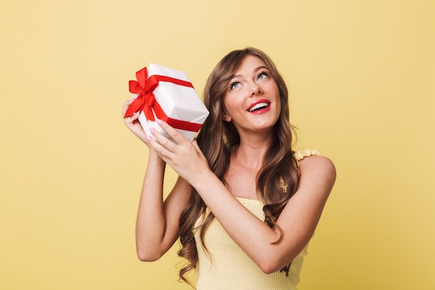 Foto di curiosa donna felice anni '20 con lunghi capelli castani sorridente e scuotendo confezione regalo per capire cosa c'è dentro, isolato su sfondo giallo