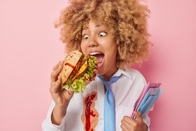 Photo of curly haired student feels very hungry after lectures\
bites tasty burger carries folders dressed in white formal shirt\
smeared with ketchup enjoys fast food isolated over pink\
background