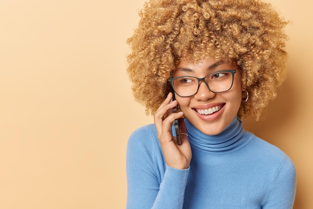 Photo of curly haired woman talks via smartphone looks\
positively aside has pleasant conversation wears transparent\
eyeglasses blue turtleneck isolated over beige background blank\
space for your text