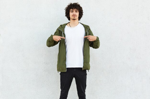 Photo of curly man points at white t shirt, shows free space for your advertisement, logo or promotional content, poses over concrete wall