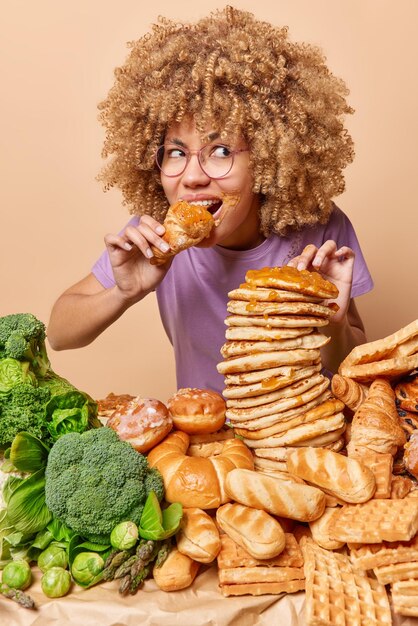 Photo of curly woman eats delicious sweet food prefers\
unhealthy nutrition tempted to eat something junk food wears\
spectacles casual t shirt feels hungry hungry female model has bad\
eating habits